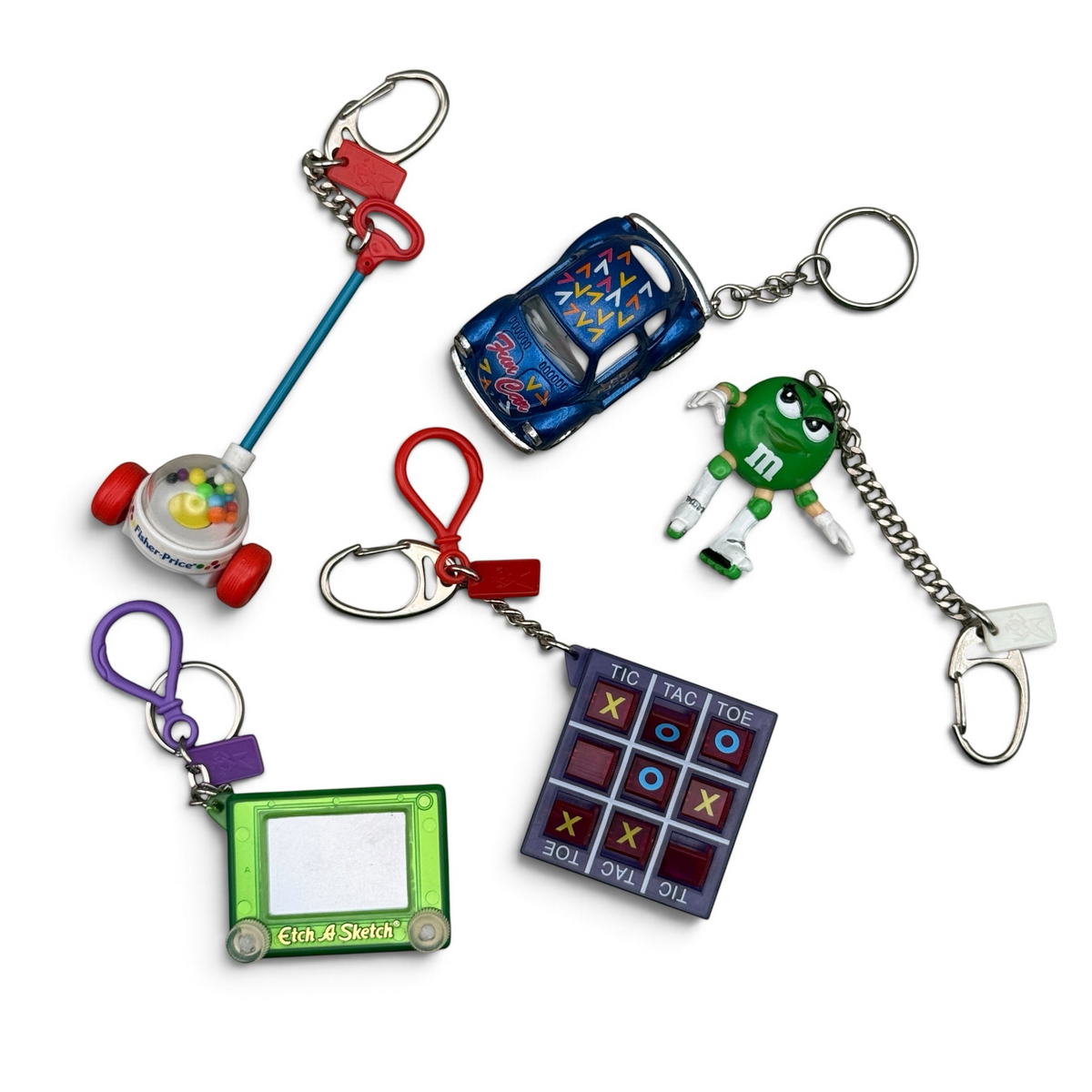 Five vintage 1980s–Y2K toy keychains: Fisher-Price Corn Popper, green Etch A Sketch, blue toy car, green M&M character, and purple Tic-Tac-Toe game, curated by BAG CRAP.
